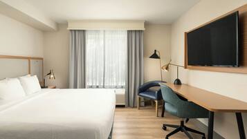 A hotel room with a large bed, a desk with a chair, a flat-screen TV, and a window with curtains.
