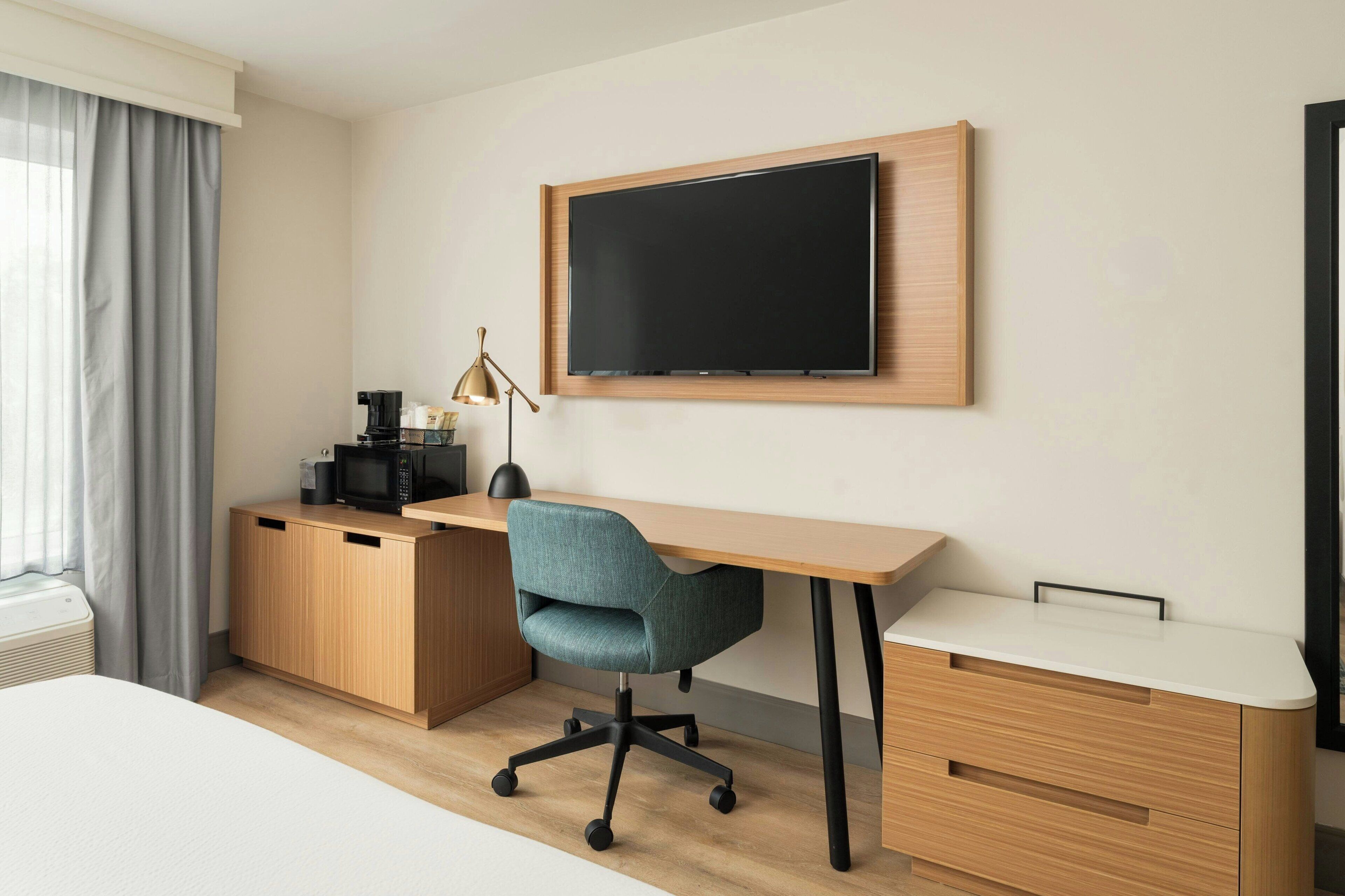 Ein modernes Hotelzimmer mit einem Holztisch, einem blaugrünen Bürostuhl, einem Flachbildfernseher, einer Kaffeemaschine und einer Lampe.