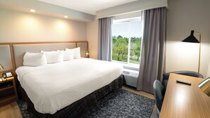A hotel room with a large bed, a desk, a chair, a nightstand, a lamp, and a view of greenery through the window. - Fairfield Inn & Suites by Marriott Jacksonville Butler Blvd (Jacksonville)