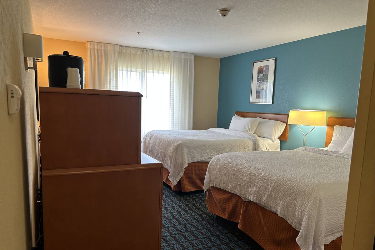 A hotel room with two beds, a refrigerator, a window with curtains, a wall-mounted picture, and a patterned carpet.