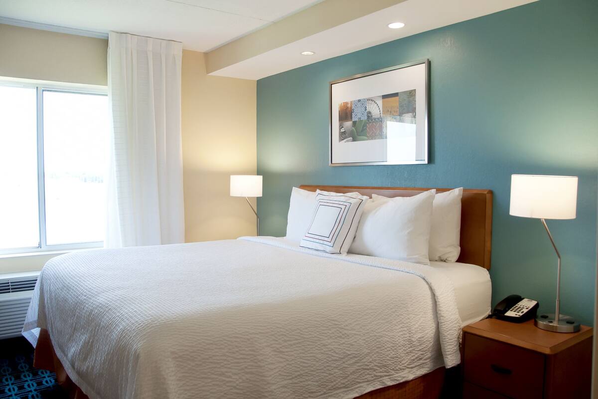 A hotel room with a large bed, a bedside table with a lamp, a phone, and a framed picture on the wall.