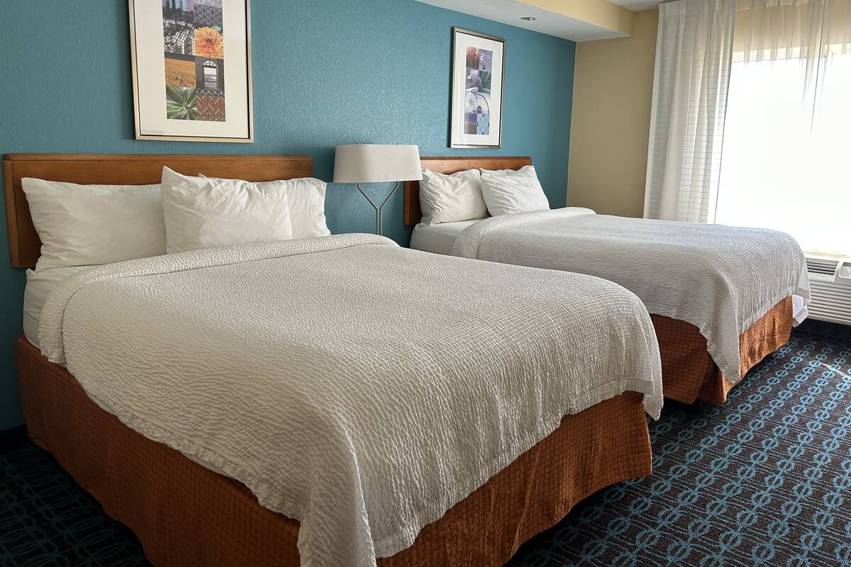 A hotel room with two beds, a headboard, a lamp, and framed pictures on the wall.