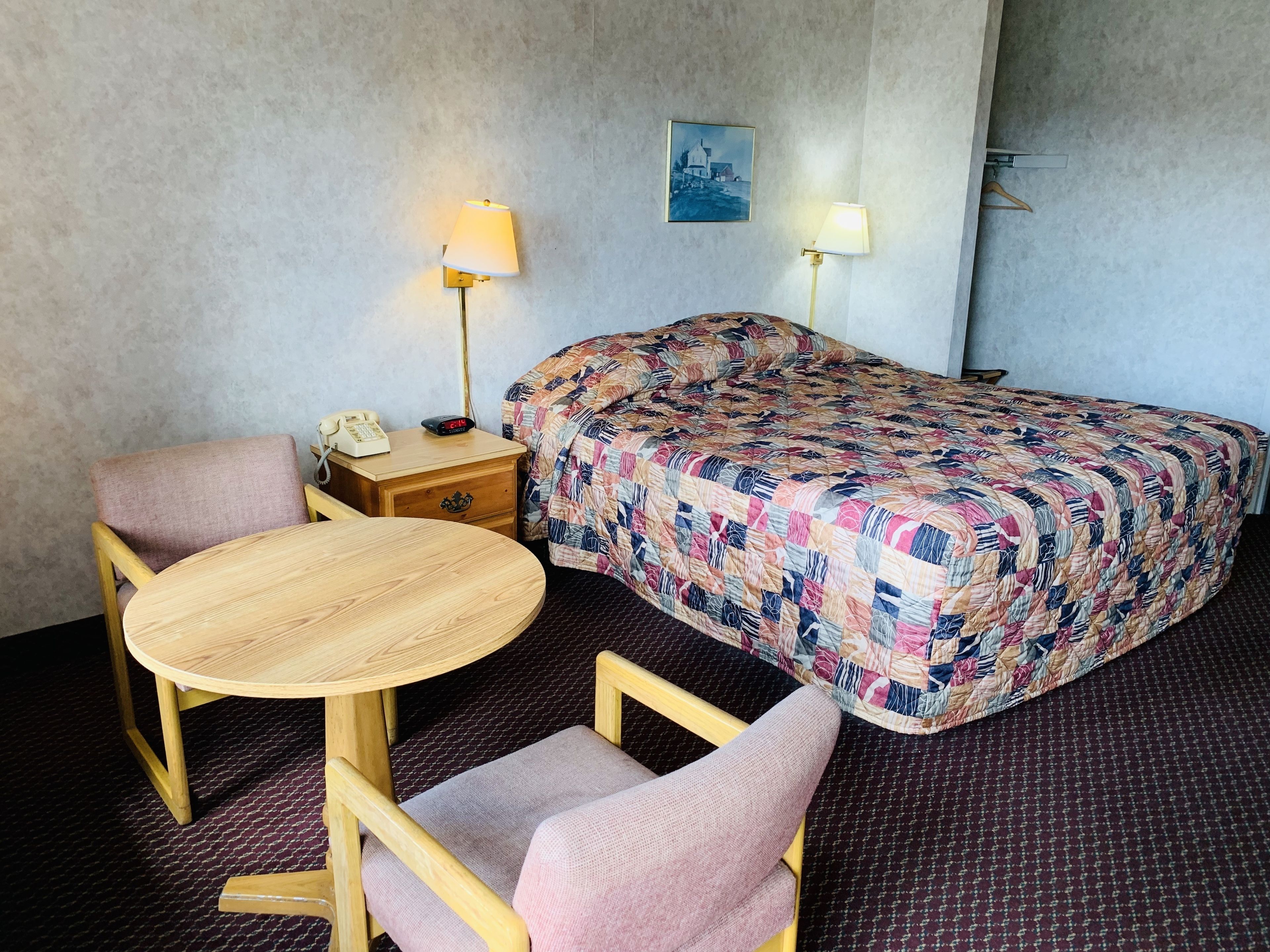 A hotel room with a bed, a round wooden table, two chairs, a nightstand with a lamp, and a painting on the wall.