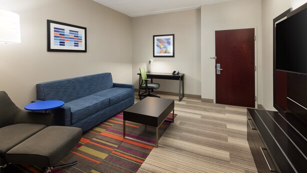 A modern hotel room with a blue sofa, a small table, a desk with a chair, a TV, and a door.