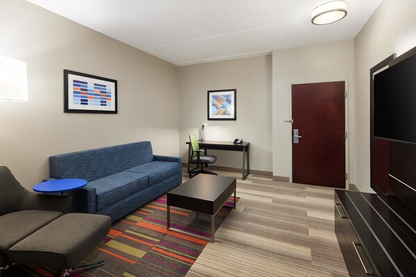 A modern hotel room with a blue sofa, a small table, a desk with a chair, a TV, and a door. - Holiday Inn Express & Suites Seymour by IHG (Seymour)