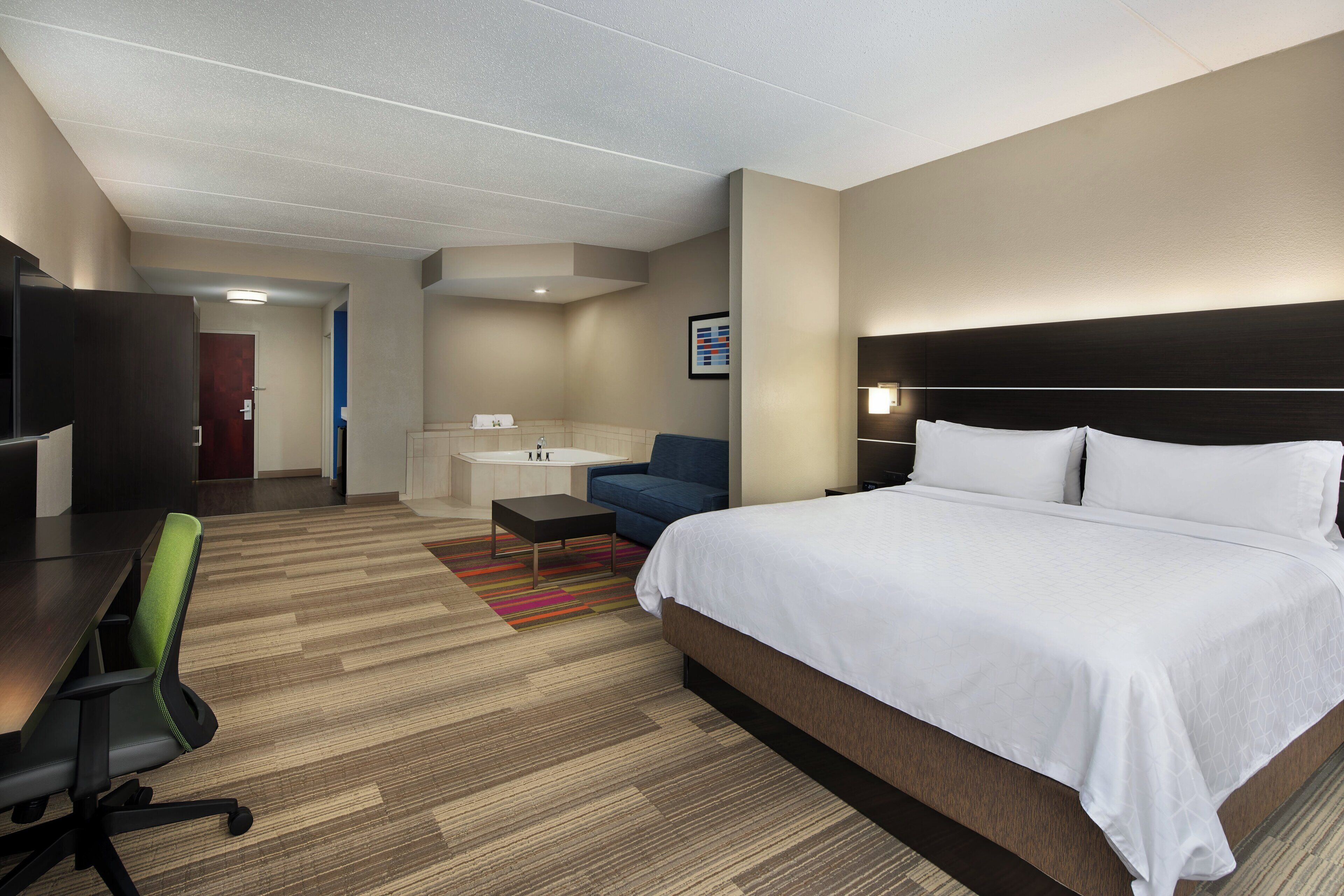 A hotel room with a large bed, a desk, a chair, a sofa, and a bathroom visible in the background.