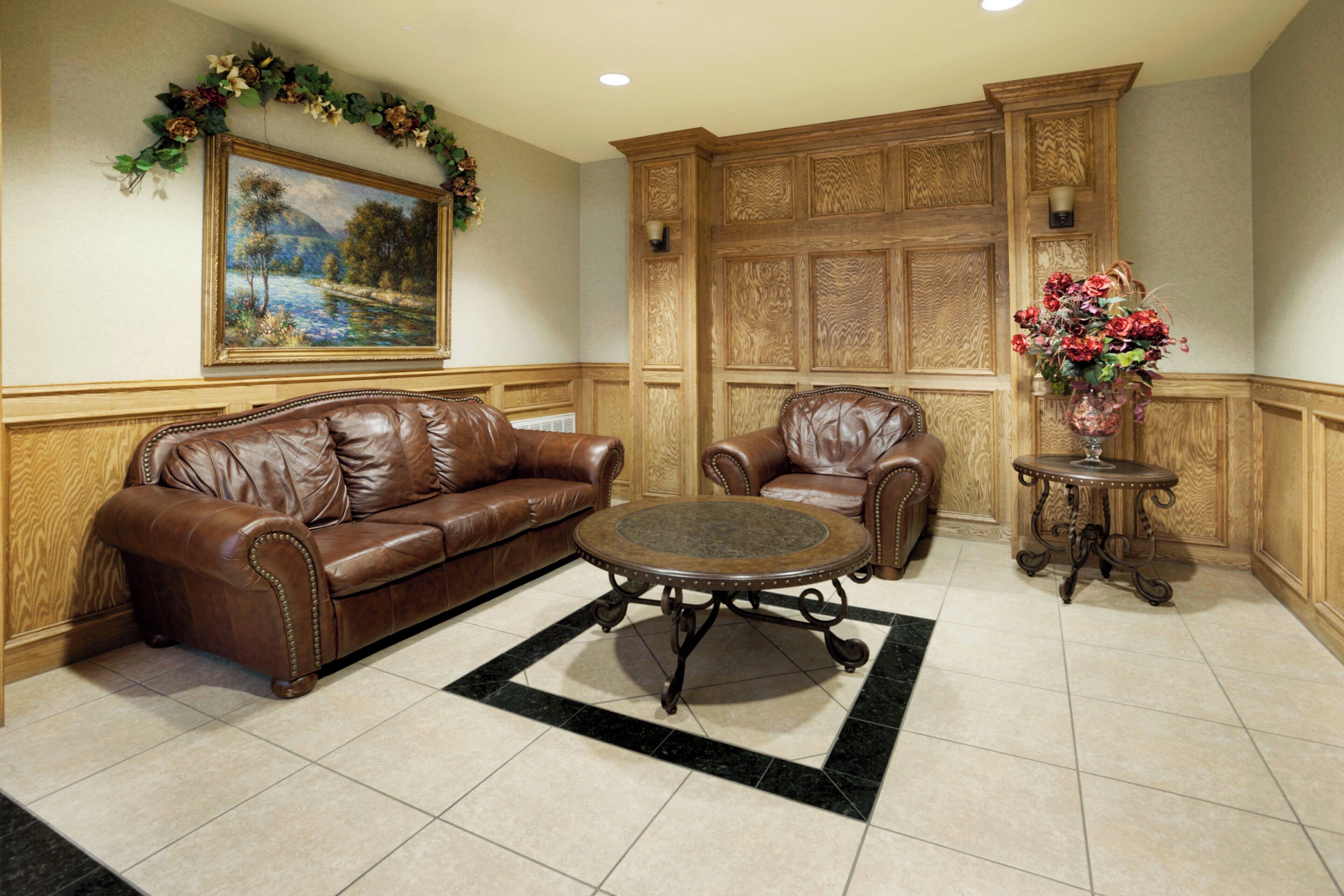 A room with a brown leather sofa, a matching armchair, a small round table, and a painting on the wall.