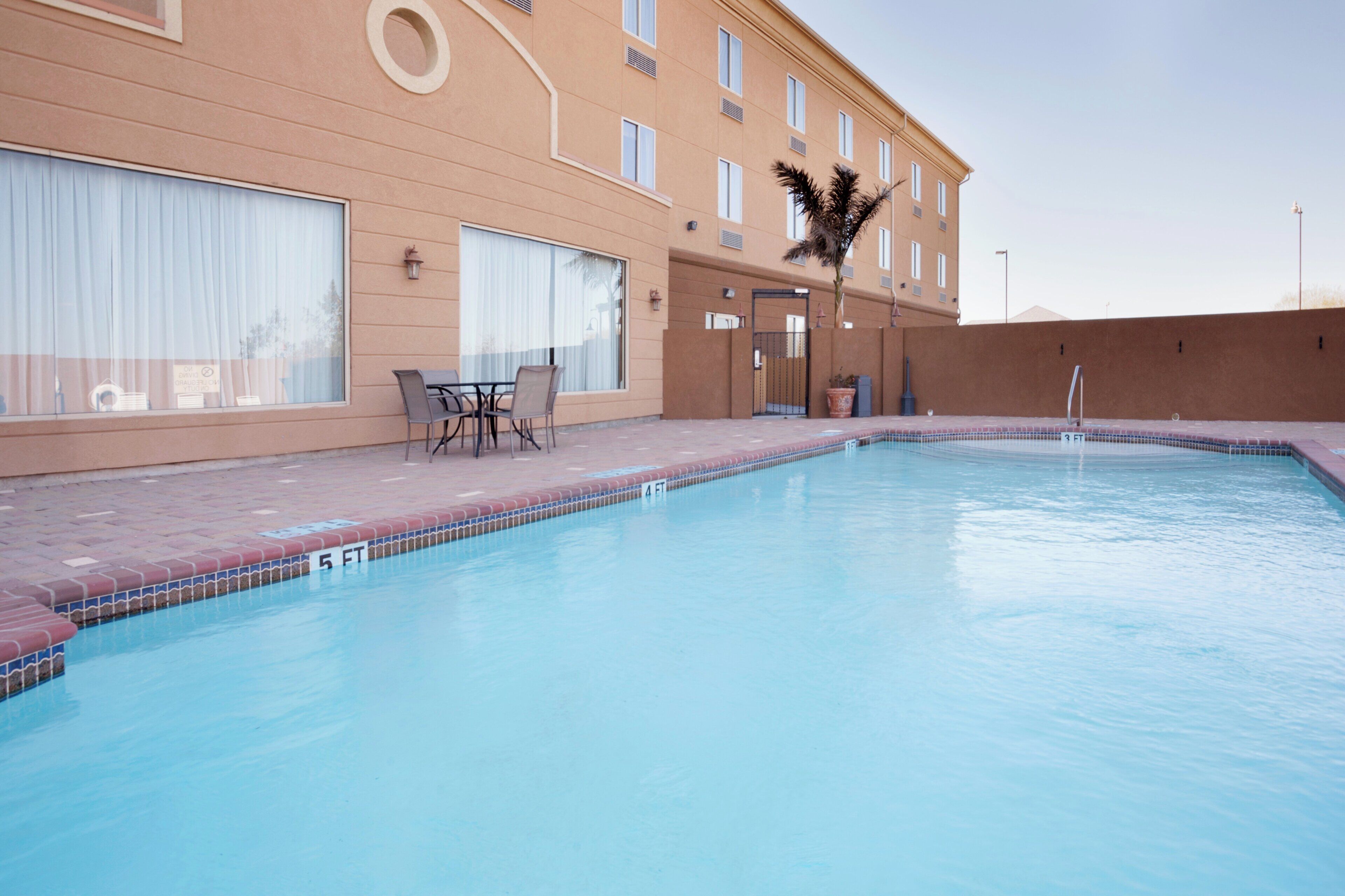 A swimming pool with a tiled edge, a lounge area with chairs, and a building with large windows and a palm tree.