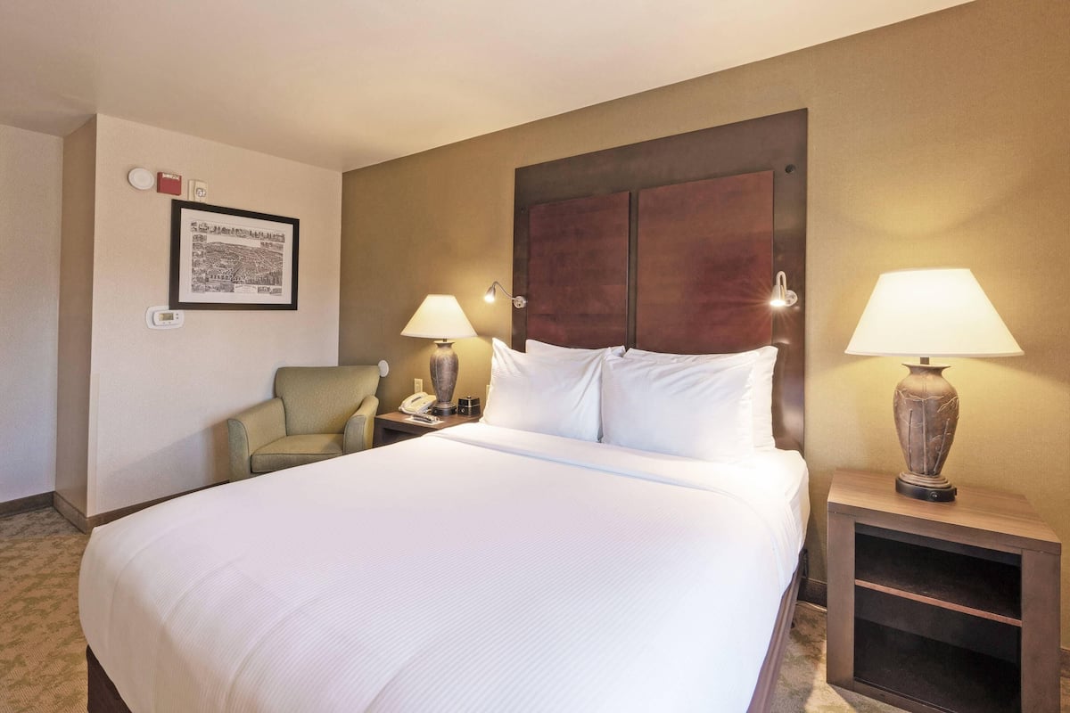 A hotel room with a large bed, two bedside tables with lamps, a chair, a framed picture on the wall, and a small shelf.