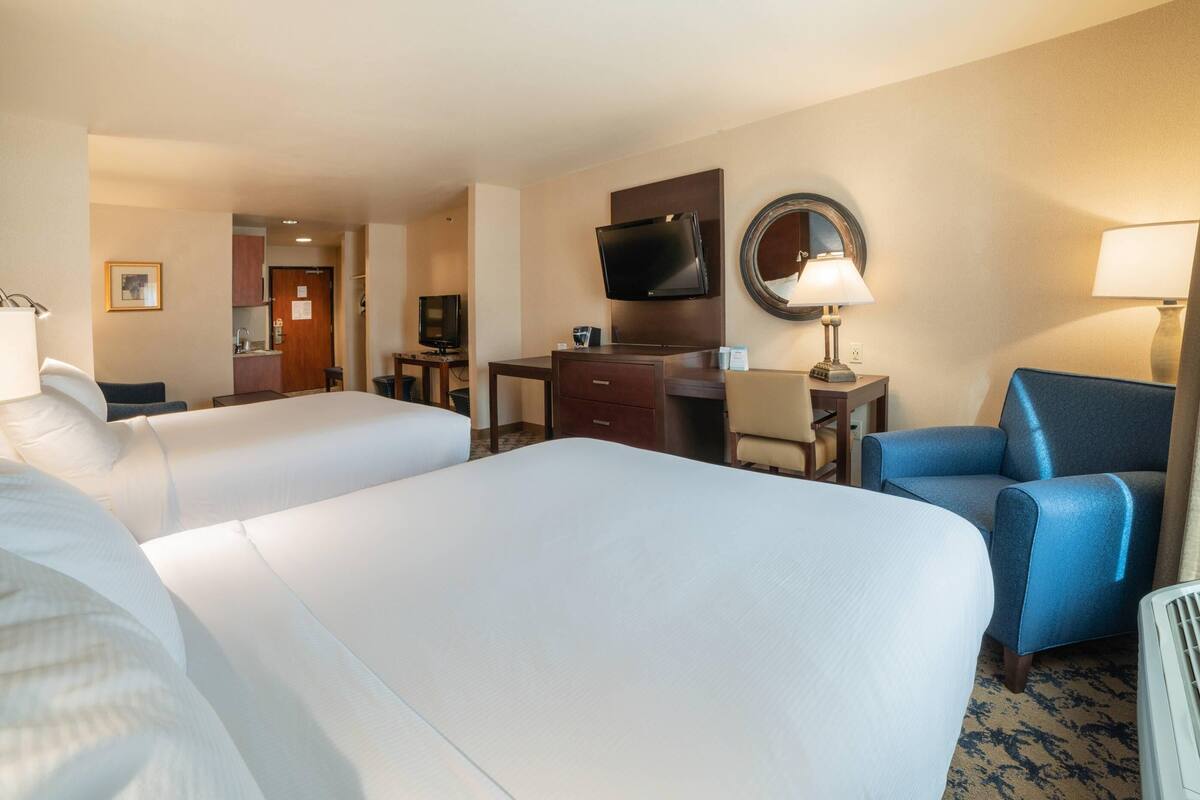 A hotel room with two beds, a desk with a flat-screen TV, a mirror, and a blue armchair.