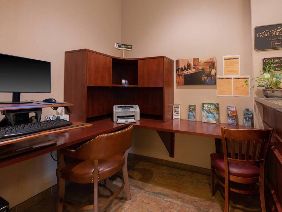 Una oficina de hotel con un escritorio, computadora, impresora y sillas. Hay carteles informativos y una planta en la pared.