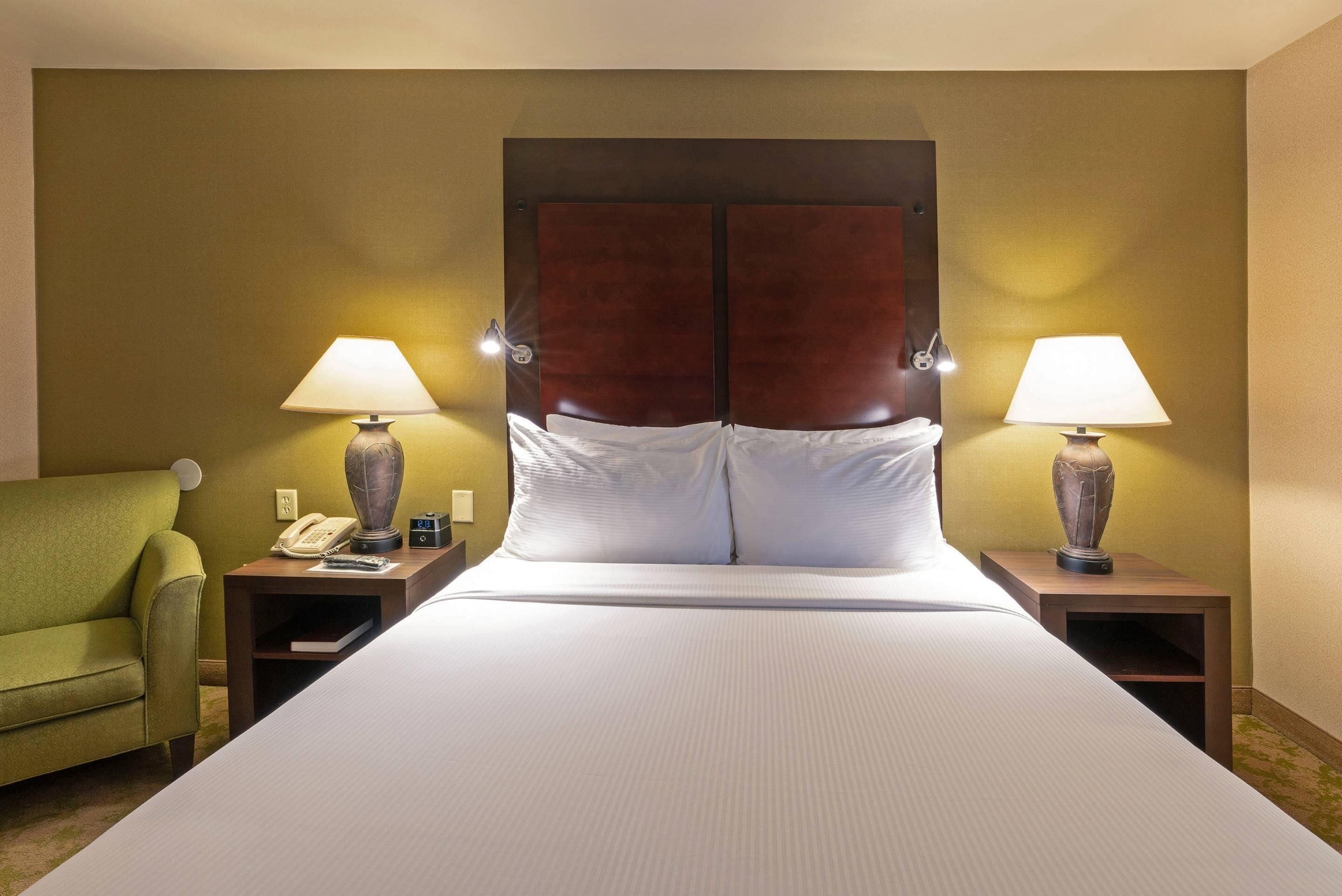 A hotel room with a large bed, two bedside lamps, a green armchair, a bedside telephone, and a small table with a lamp.