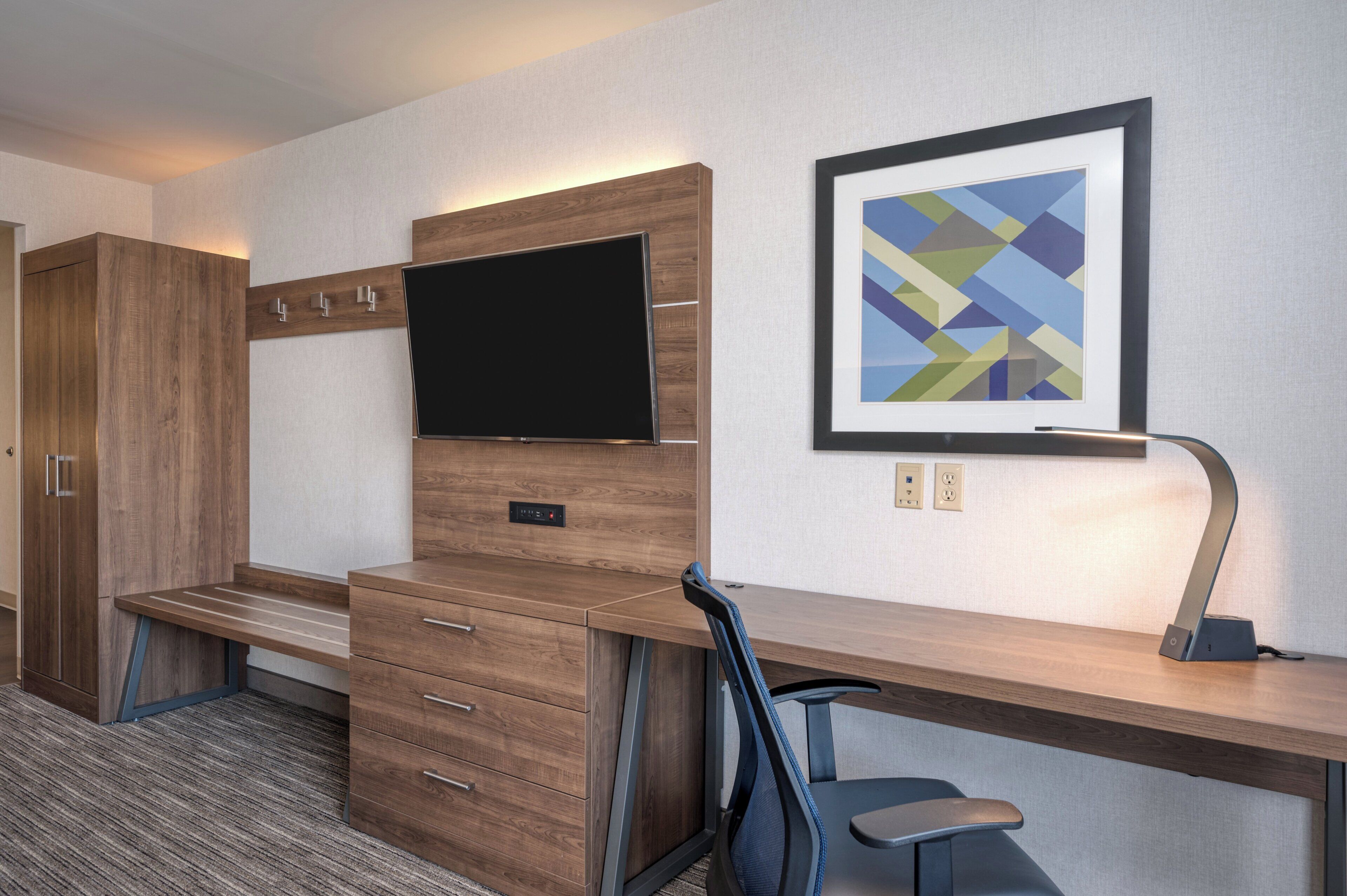 A hotel room with a wooden desk, a flat-screen TV, a framed abstract painting, and a desk lamp.