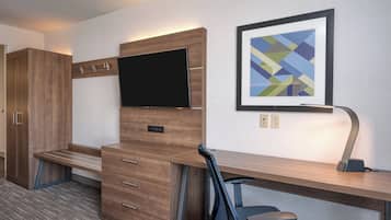 A hotel room with a wooden desk, a flat-screen TV, a framed abstract painting, and a desk lamp.