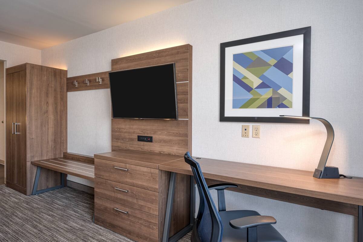 A hotel room with a wooden desk, a flat-screen TV, a framed abstract artwork, and a bench.
