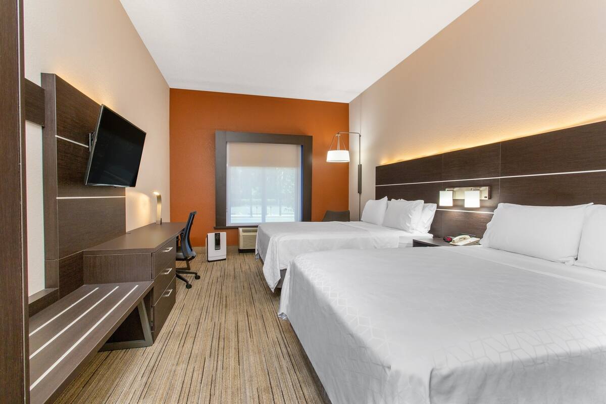 A hotel room with two beds, a desk, a flat-screen TV, a desk lamp, and a window with blinds.