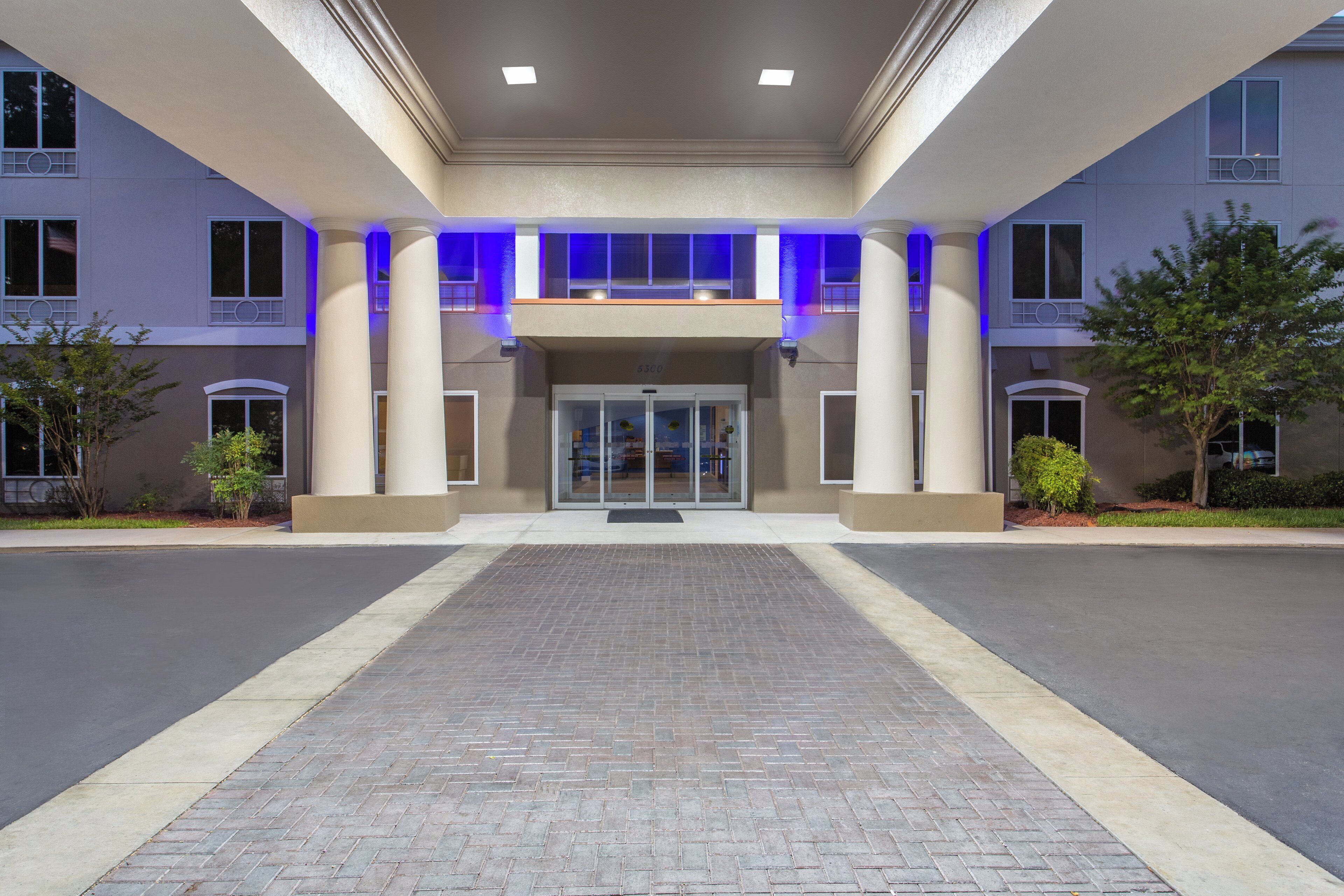 A hotel entrance with a brick pathway leading to a glass door entrance, flanked by columns and trees.