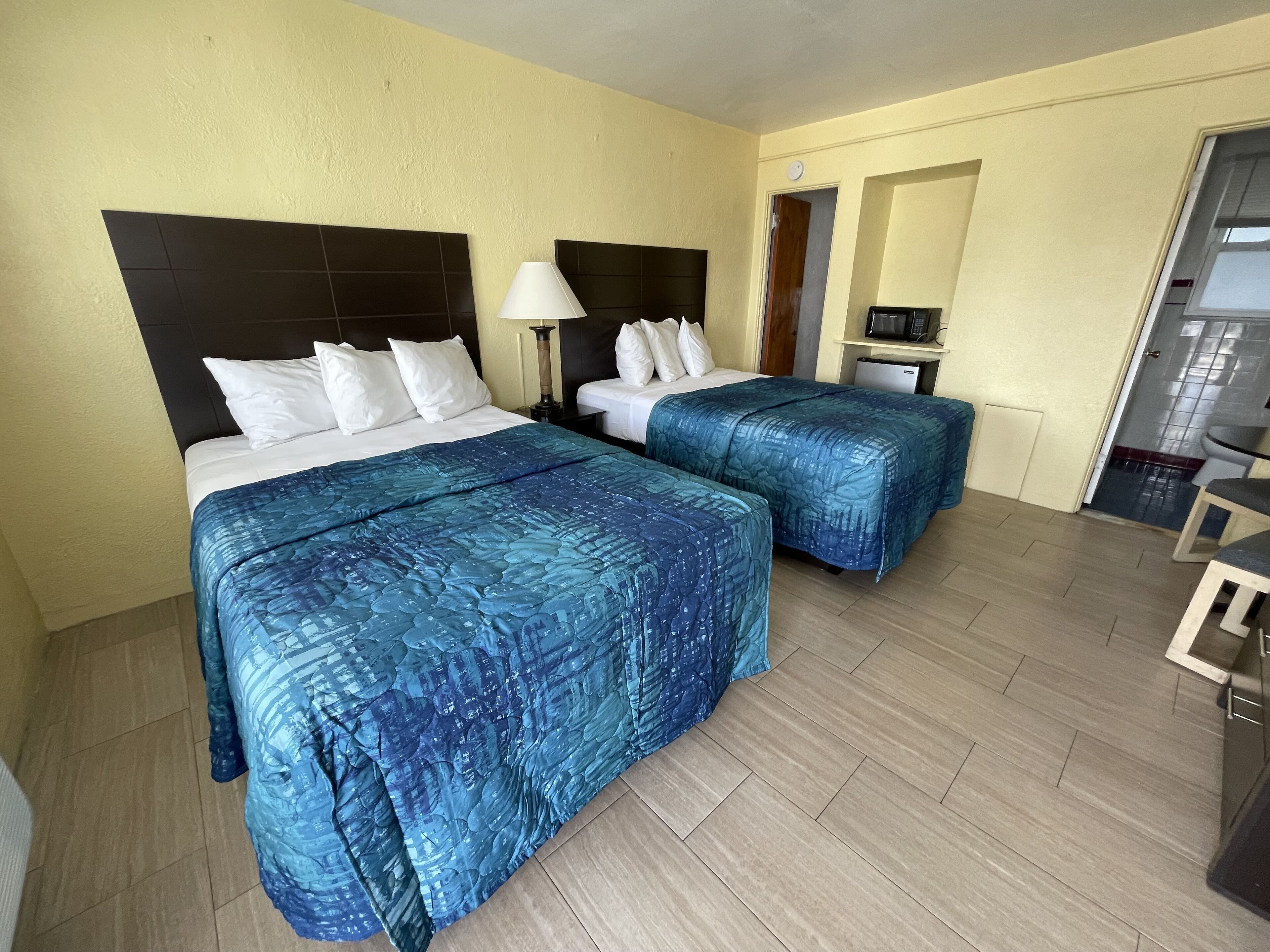 A hotel room with two beds, each with a headboard, white bedding, and blue patterned bedspreads. There is a small desk with a lamp, a microwave, and a refrigerator in the corner. The room has wooden flooring and light-colored walls.