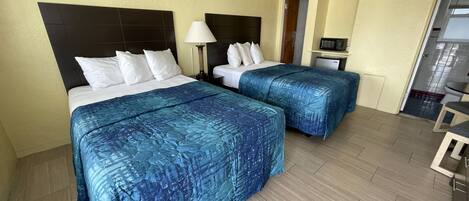 A hotel room with two beds, each with a headboard, white bedding, and blue patterned bedspreads. There is a small desk with a lamp, a microwave, and a refrigerator in the corner. The room has wooden flooring and light-colored walls.