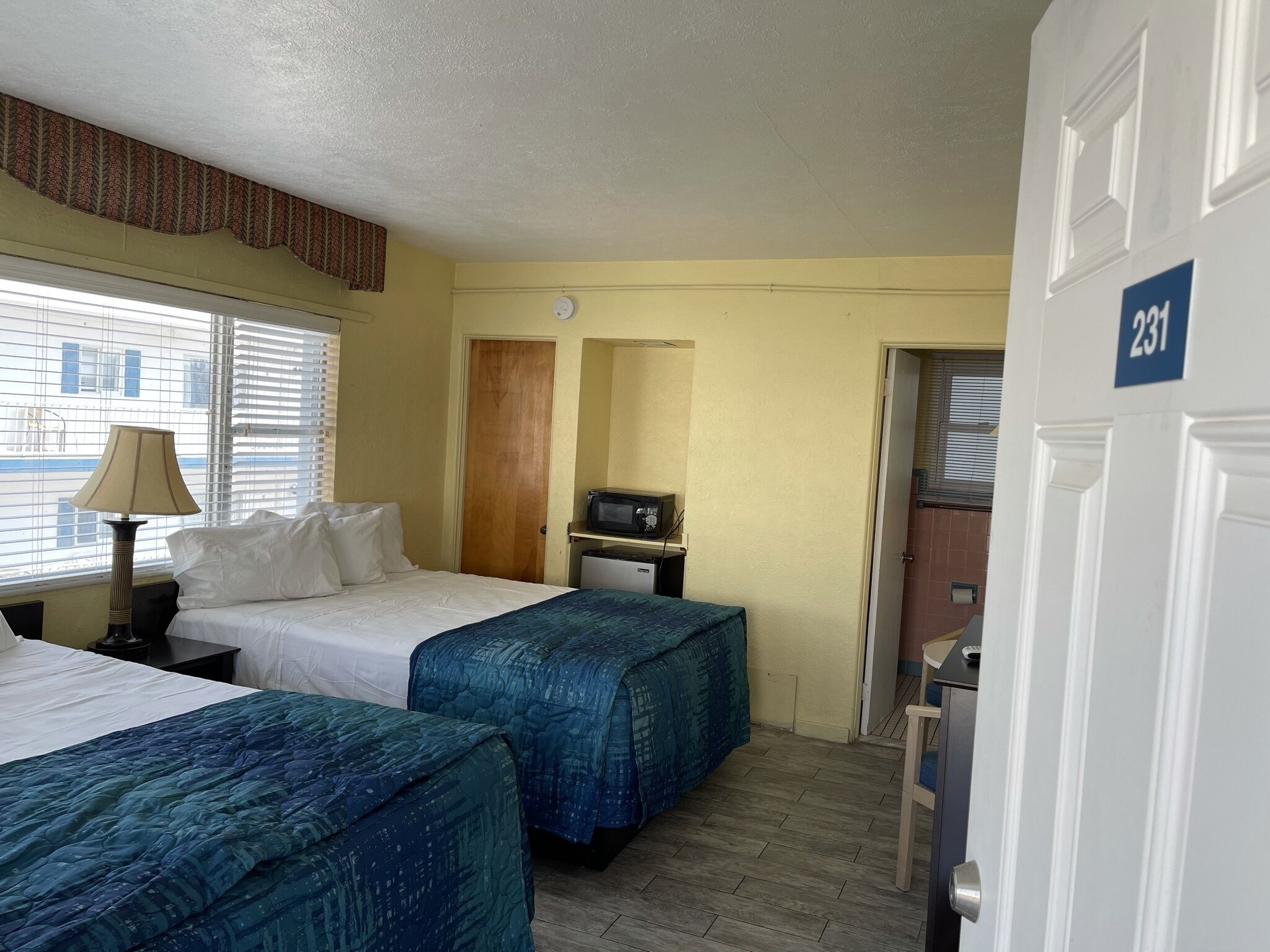 A hotel room with two beds, a desk with a lamp, a microwave, and a door marked with the number 231.