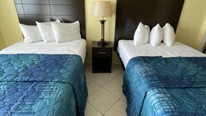 A hotel room with two beds, each with a headboard, white bedding, and blue patterned bedspreads. There is a nightstand with a lamp between the beds, and two paintings of boats on the wall. - Sandy Shores Resort Motel (North Wildwood)