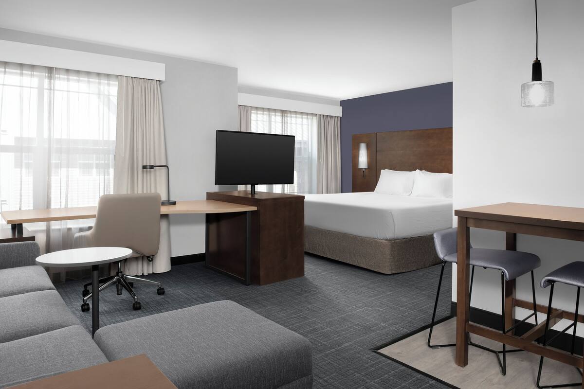 A hotel room with a bed, a desk with a TV, a sofa, and a small bar area.