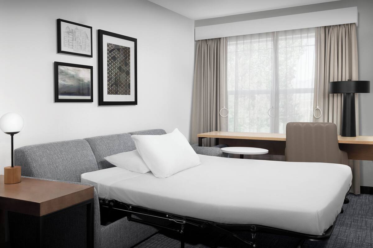 A hotel room with a bed, a desk, a chair, a lamp, and framed pictures on the wall.