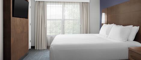 Ein Hotelzimmer mit einem großen Bett, einem flachbildfernseher an einer Holzverkleidung, einem Fenster mit transparenten Vorhängen und einem Nachttisch mit einer Lampe.