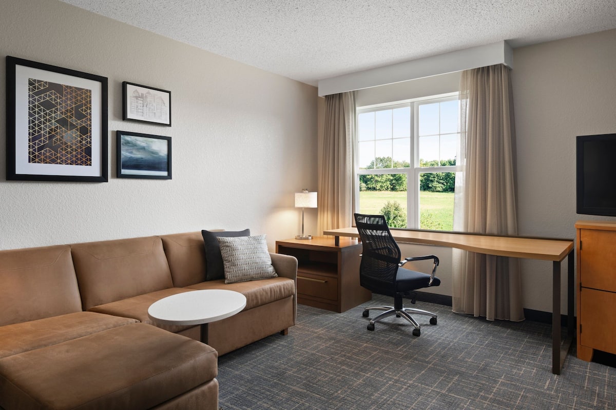 A hotel room with a brown leather sofa, a small round table, a desk with a chair, a TV, and framed pictures on the wall.