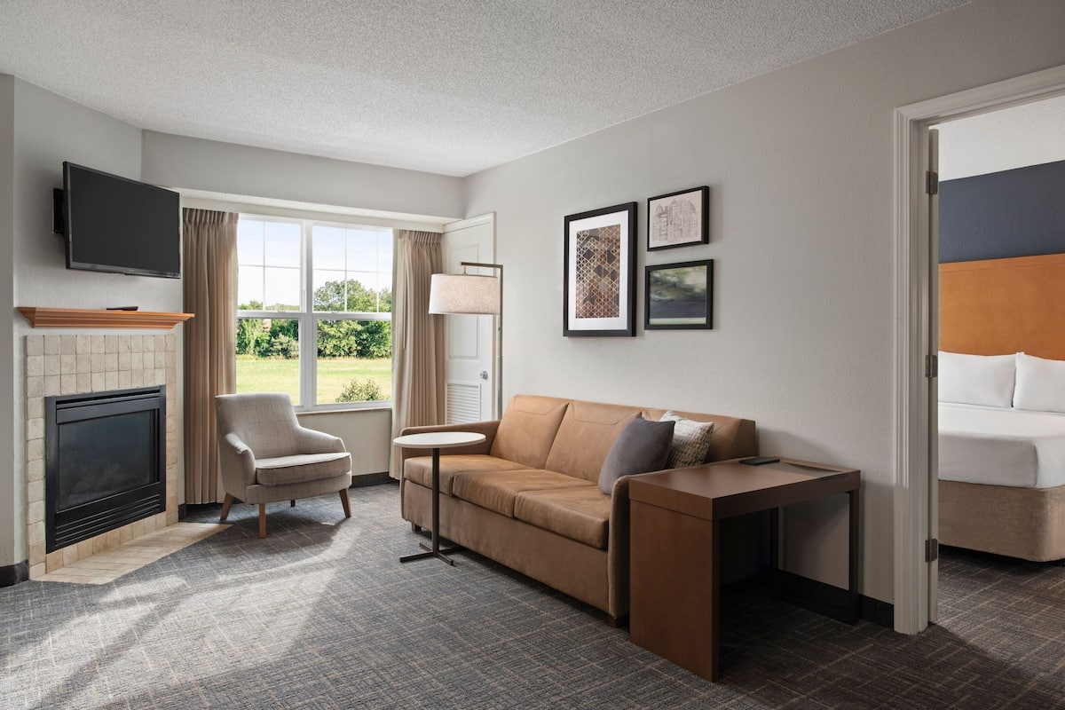 A hotel room with a fireplace, a sofa, a small table, a TV mounted on the wall, and framed pictures on the wall.