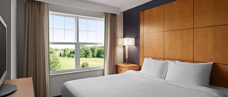 A hotel room with a large bed, a wooden headboard, a bedside table with a lamp, a TV, and a window with curtains.