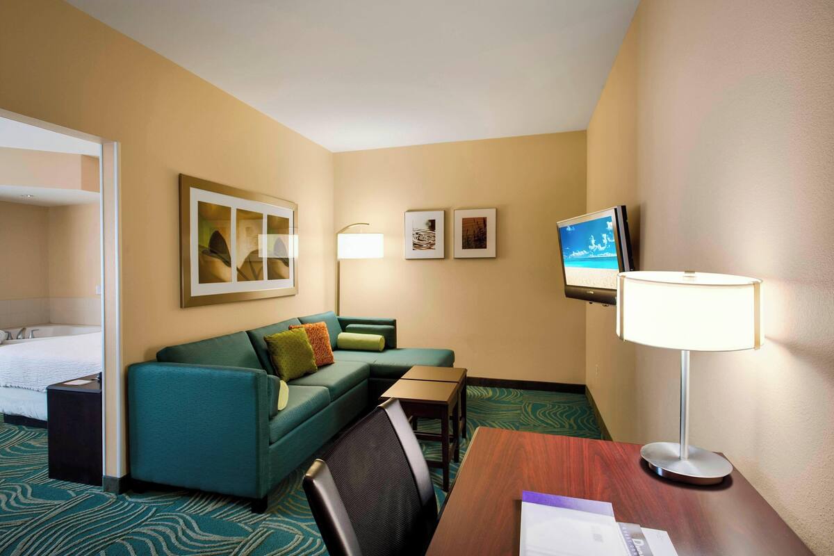 Habitación de hotel con cama, sofá verde, escritorio, silla, televisión y lámpara.