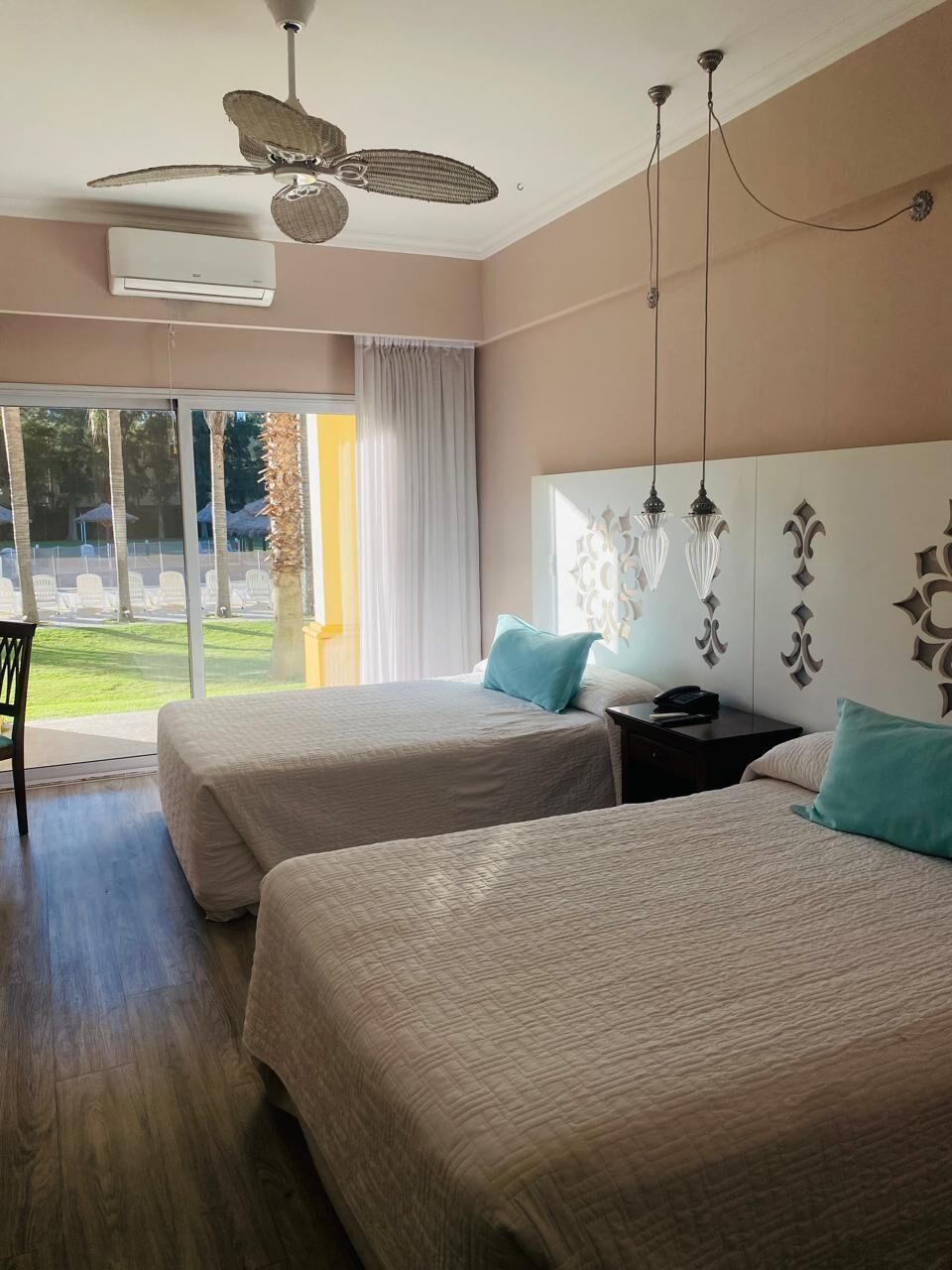 Habitación de hotel con dos camas, ventilador de techo, equipo de aire acondicionado, mesita de noche y vista a un patio.