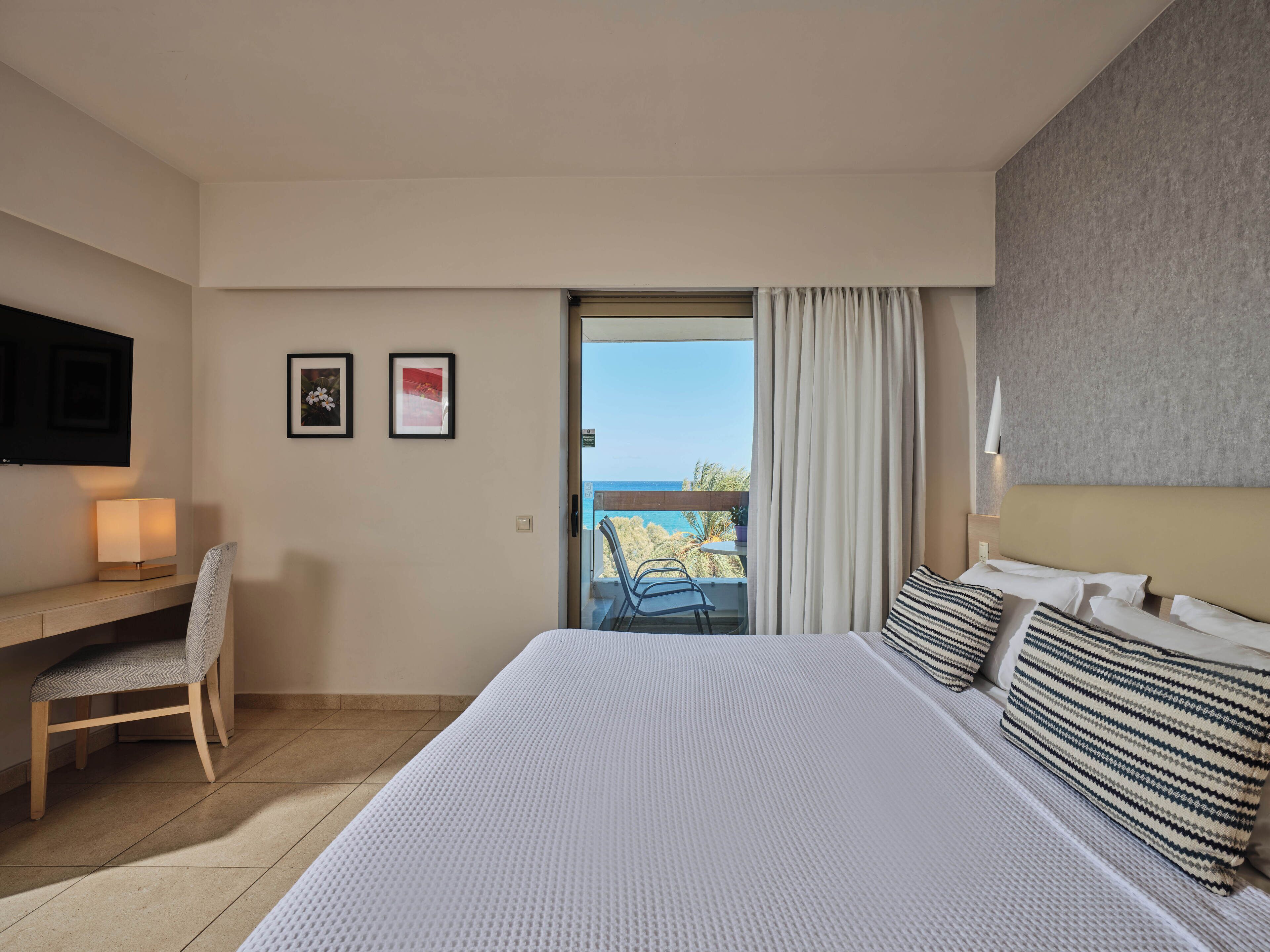 Habitación de hotel con una cama grande, un escritorio con silla, un televisor montado en la pared y un balcón con vistas al mar.