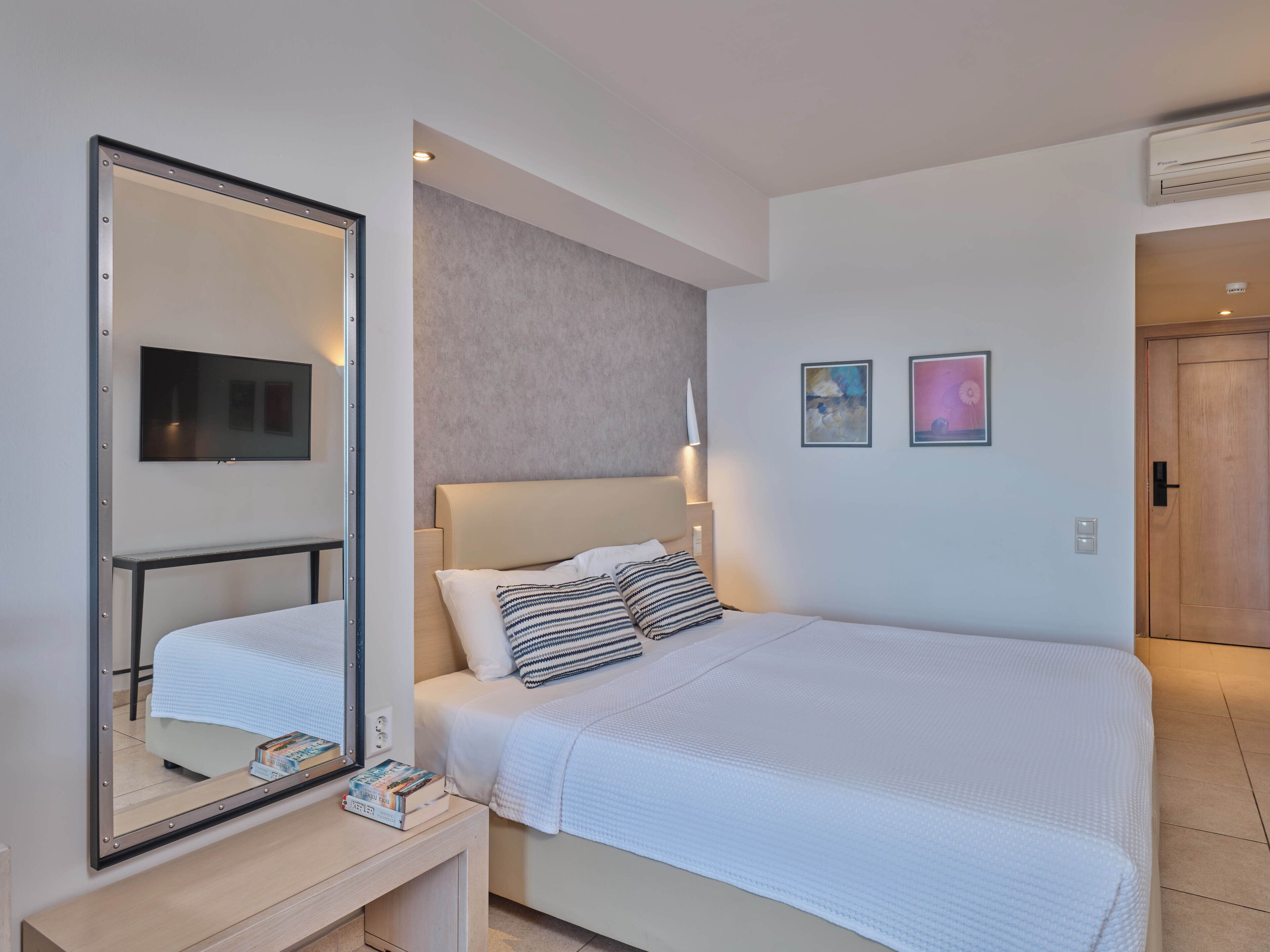 A modern hotel room with a large bed, a bedside table with books, a mirror, and a wall-mounted TV.