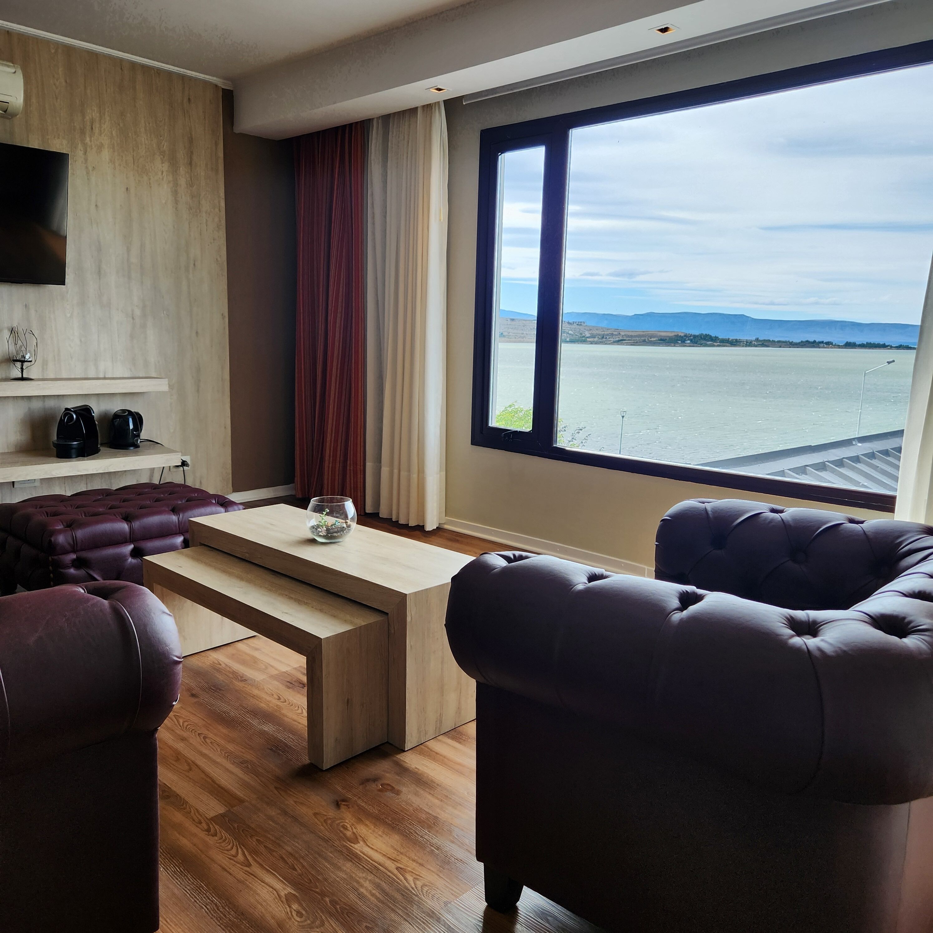 A modern living room with a large window, a flat-screen TV, a wooden coffee table, and dark leather sofas.