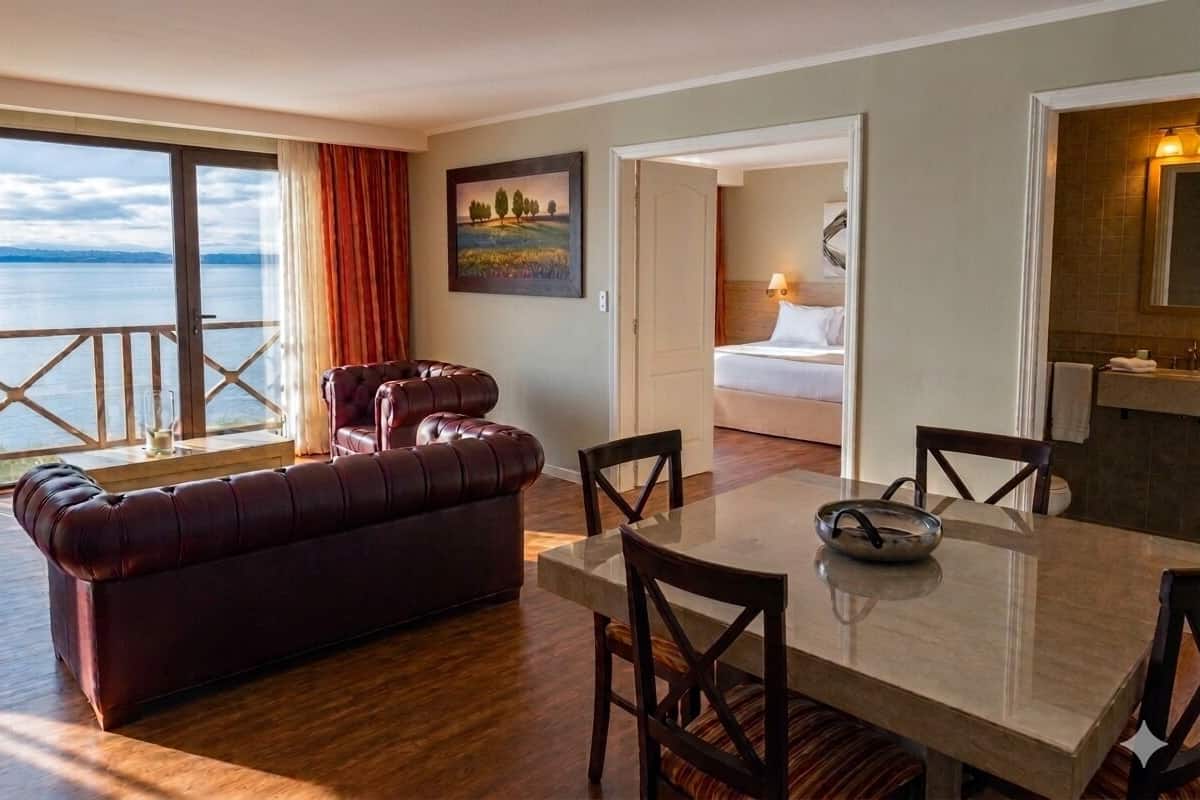 A room with a large window, a sofa, a dining table with chairs, and a bed.