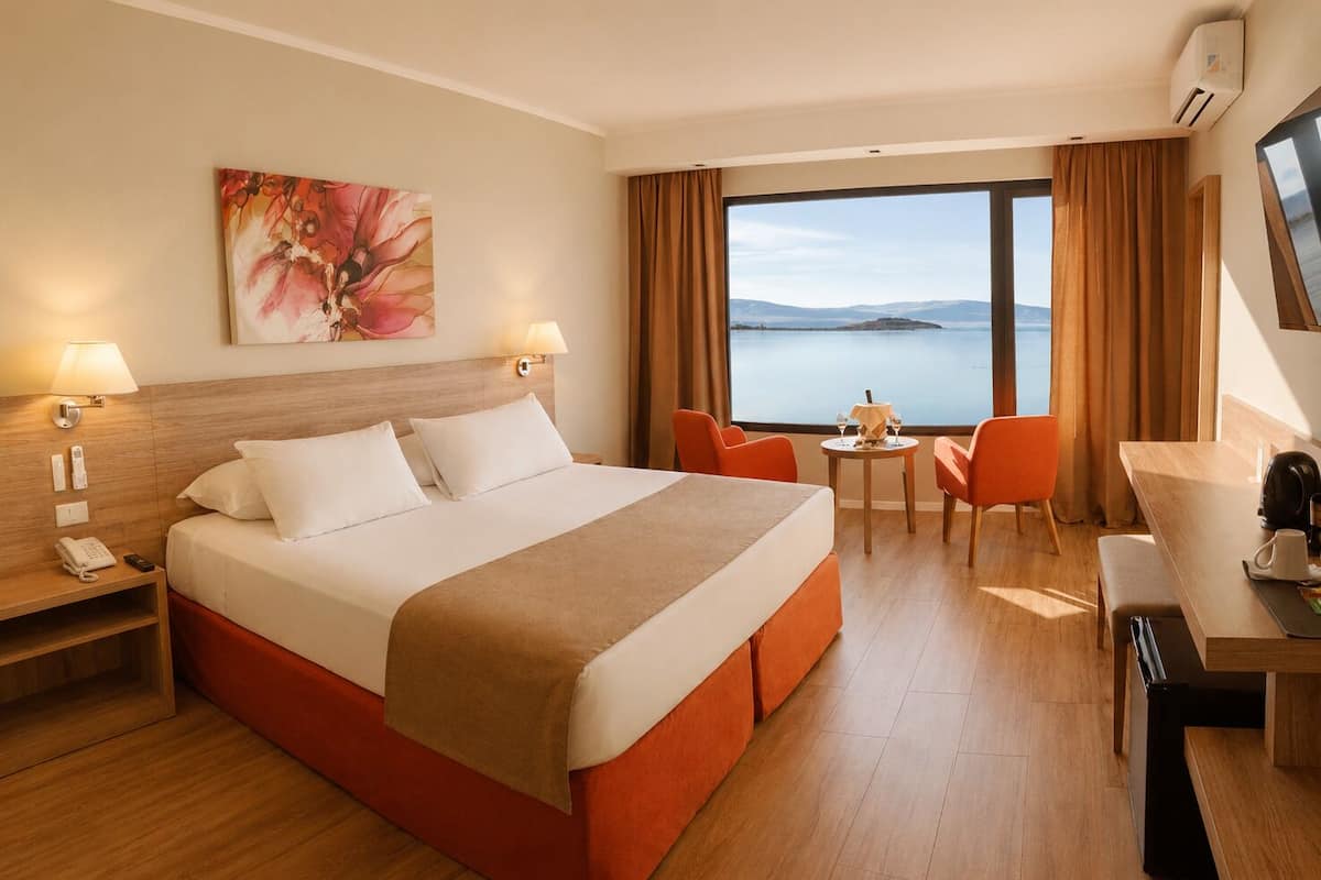 A hotel room with a bed, a desk, a chair, a table, a lamp, a phone, and a painting.
