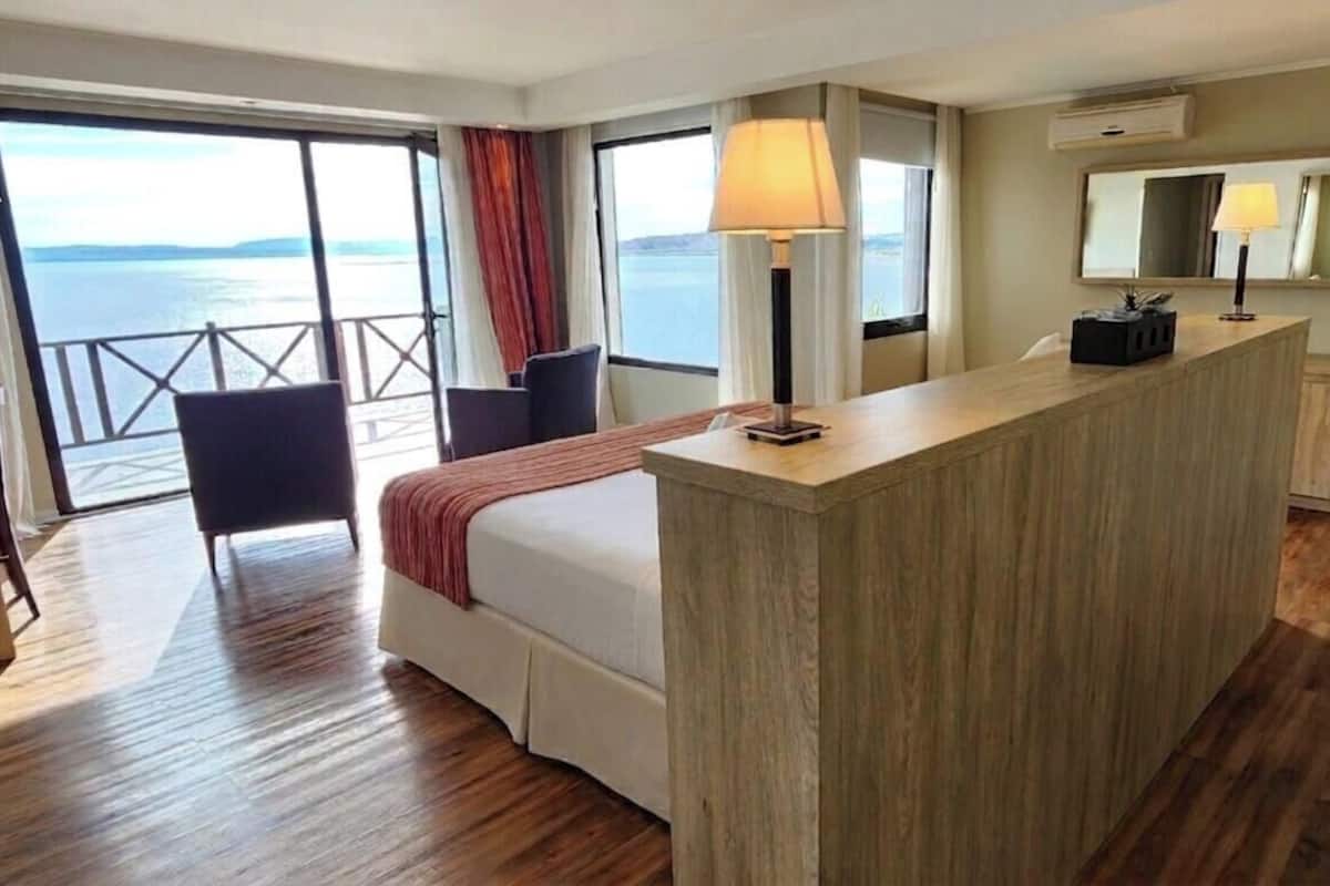 A hotel room with a bed, a desk, a chair, a large window with a view of the sea, and a balcony.