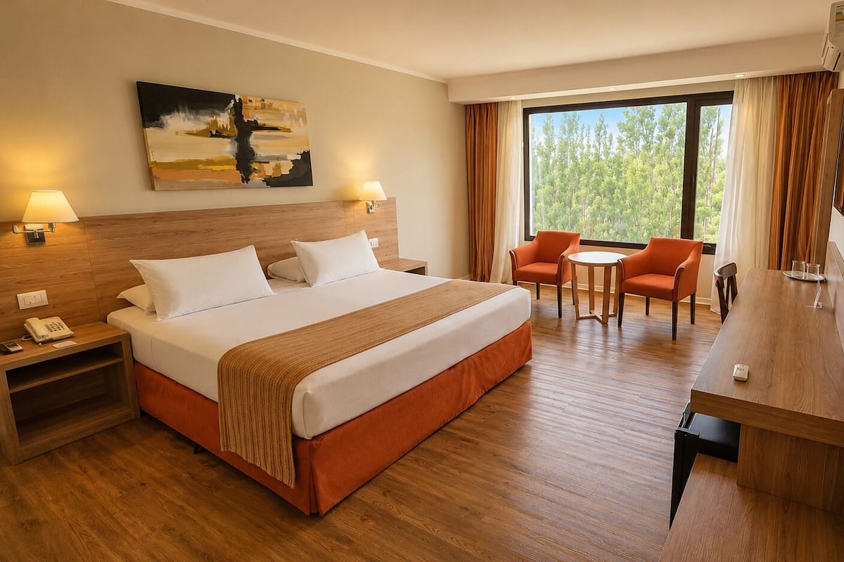 A hotel room with a bed, bedside tables, a desk, two chairs, a TV, and a large window with curtains.