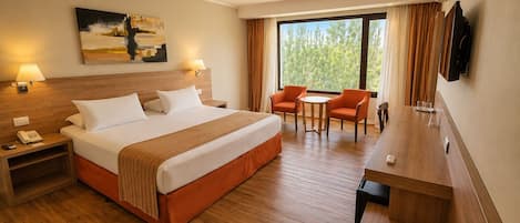 Habitación de hotel con cama, mesitas de noche, escritorio, dos sillas, televisor y un amplio ventanal con cortinas.
