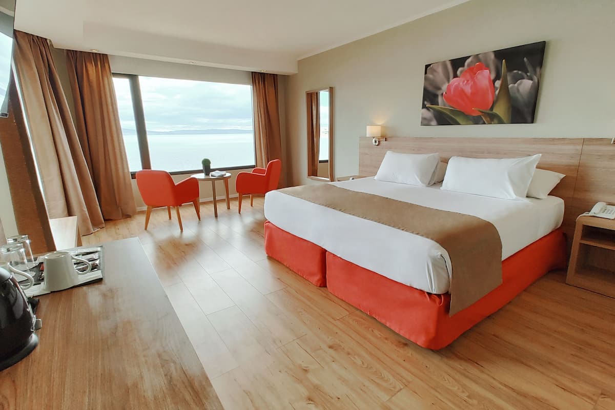 A hotel room with a large bed, a desk with a lamp, a chair, and a view of the sea through the window.