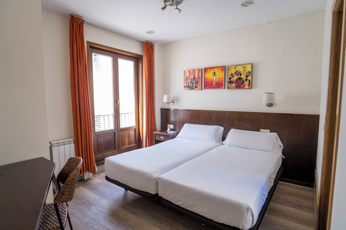 Habitació d'hotel amb dos llits, un capçal de fusta, una taula de menjador i una finestra amb cortines.