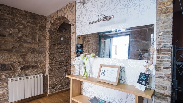 Una habitación con paredes de piedra, suelo de madera, un radiador, un espejo, una estantería con un certificado enmarcado y un florero con ramas.
