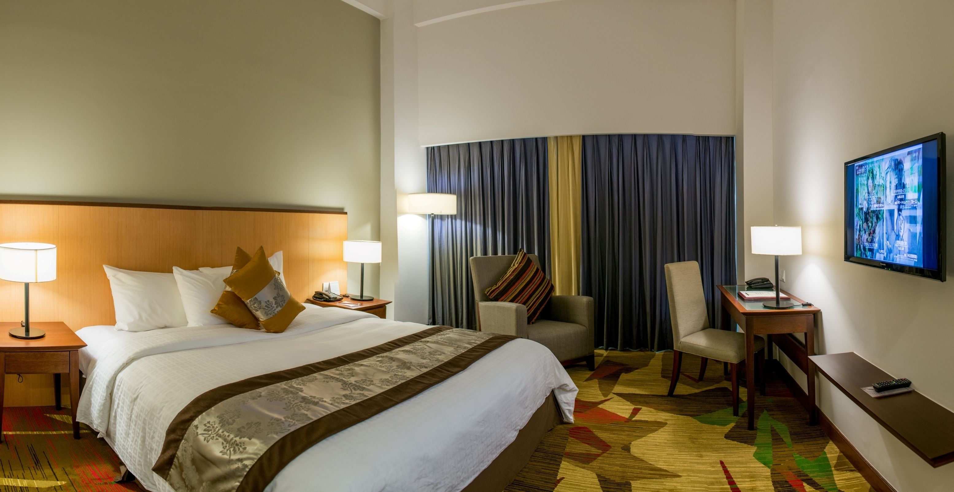 Une chambre d’hôtel avec un grand lit, deux tables de chevet avec des lampes, une chaise, un bureau et un téléviseur fixé au mur.