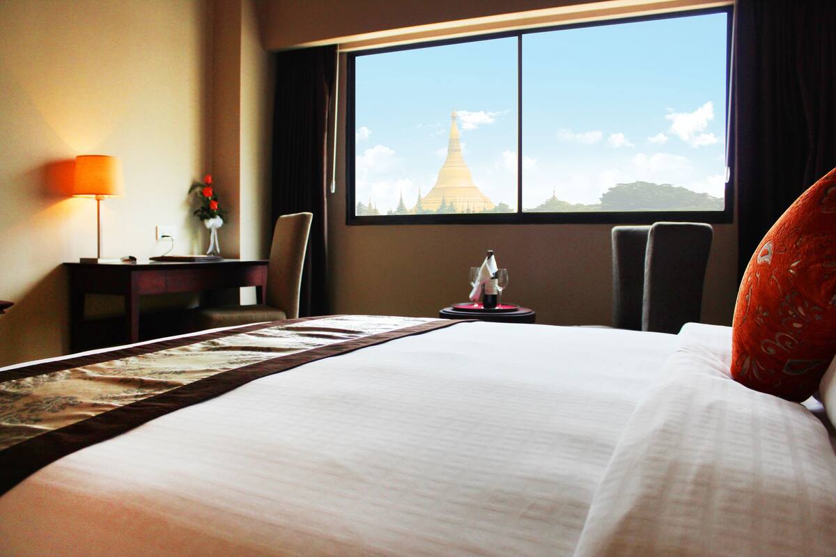 A hotel room with a large bed, a desk with a lamp, a chair, and a view of a temple through the window.