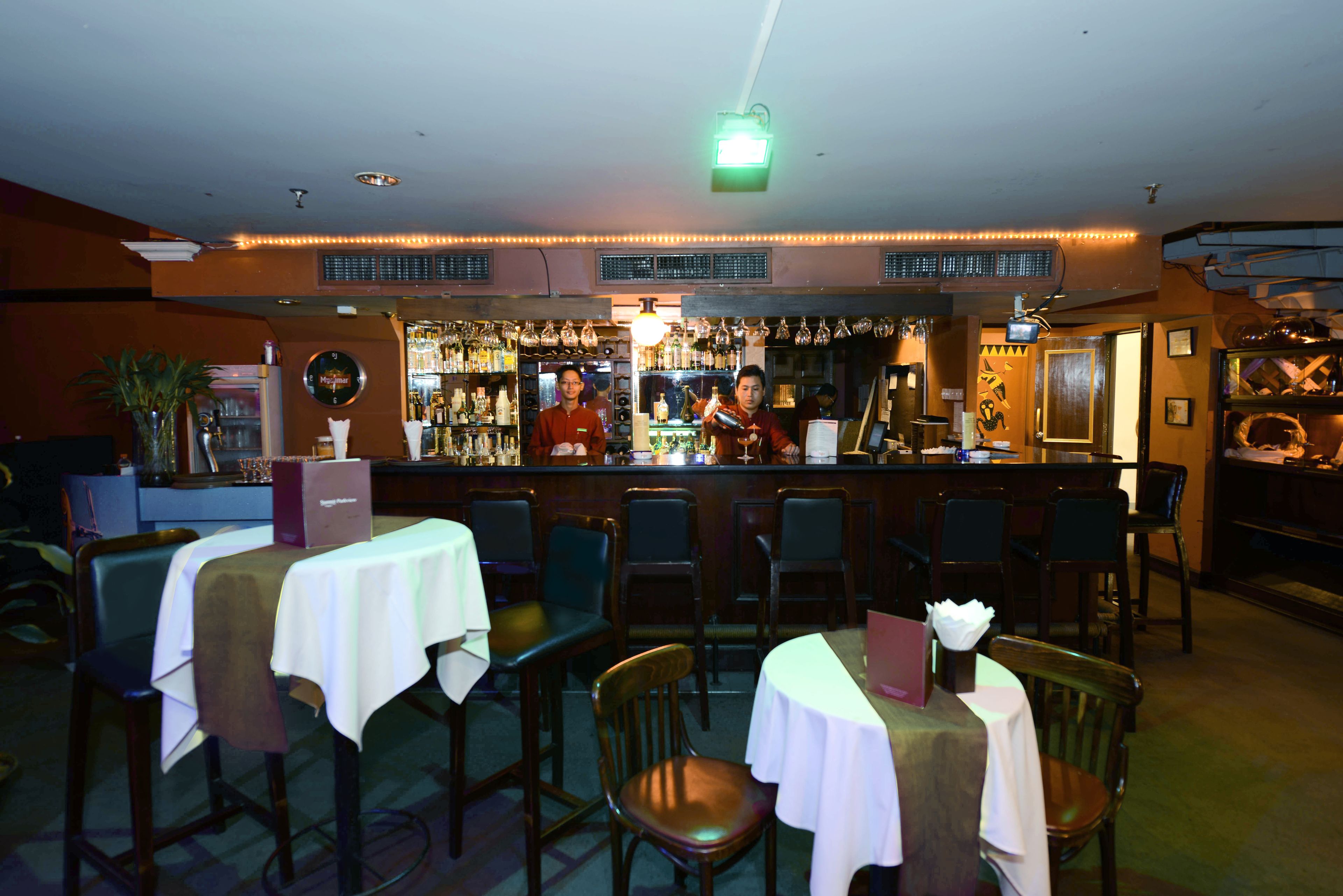 En bar med borde, stole, en bardisk med bartender og hylder fyldt med flasker.