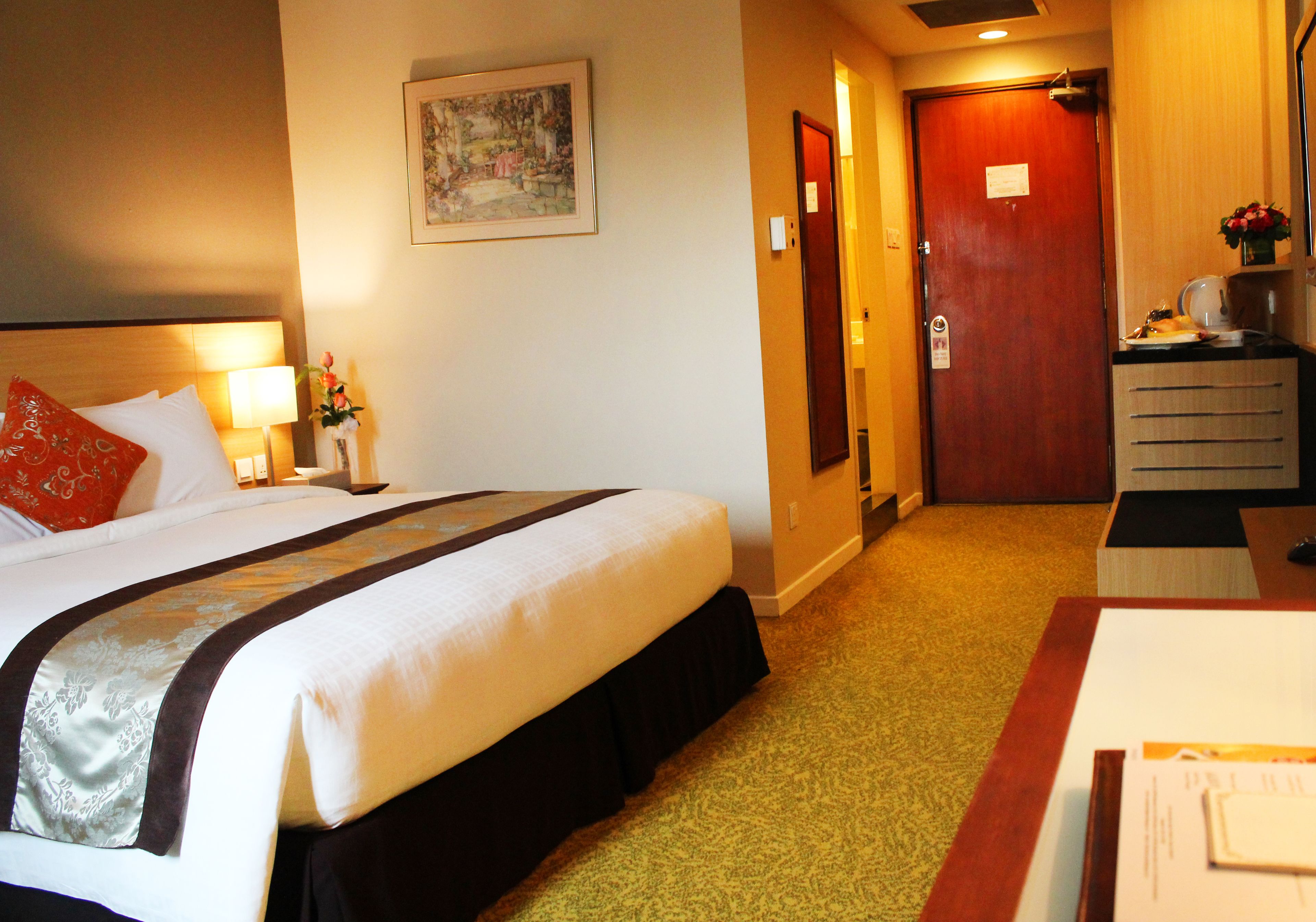 A hotel room with a bed, a desk, a chair, a lamp, a painting on the wall, and a door leading to another room.