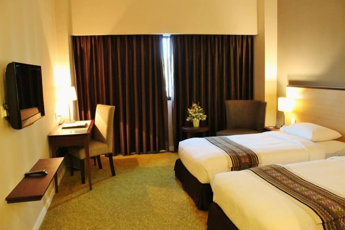 Habitación de hotel con dos camas, un escritorio con silla, televisión y una mesita con un jarrón de flores.