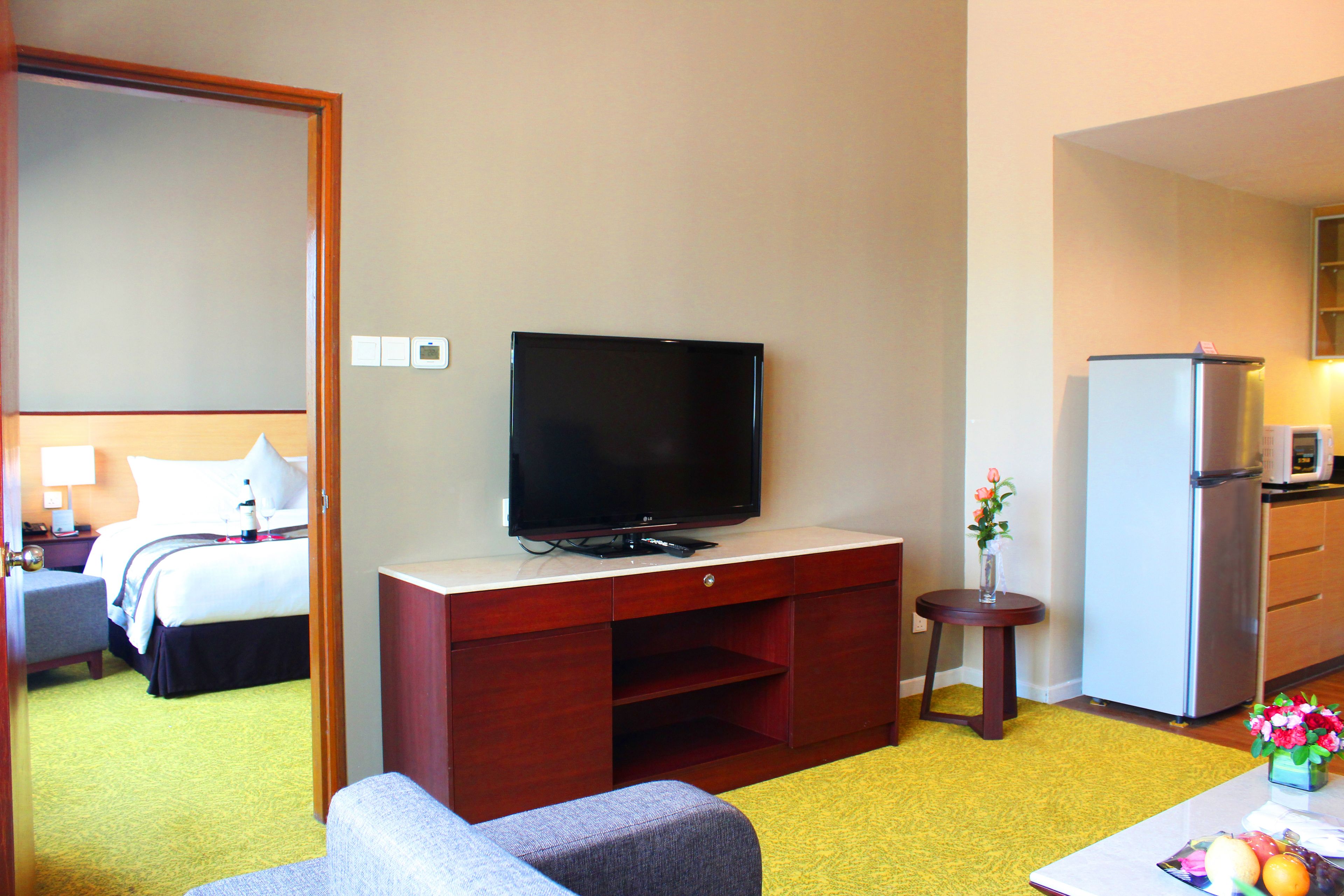 A hotel room with a bed, a TV on a wooden cabinet, a small table with a vase of flowers, and a kitchen area with a refrigerator and microwave.